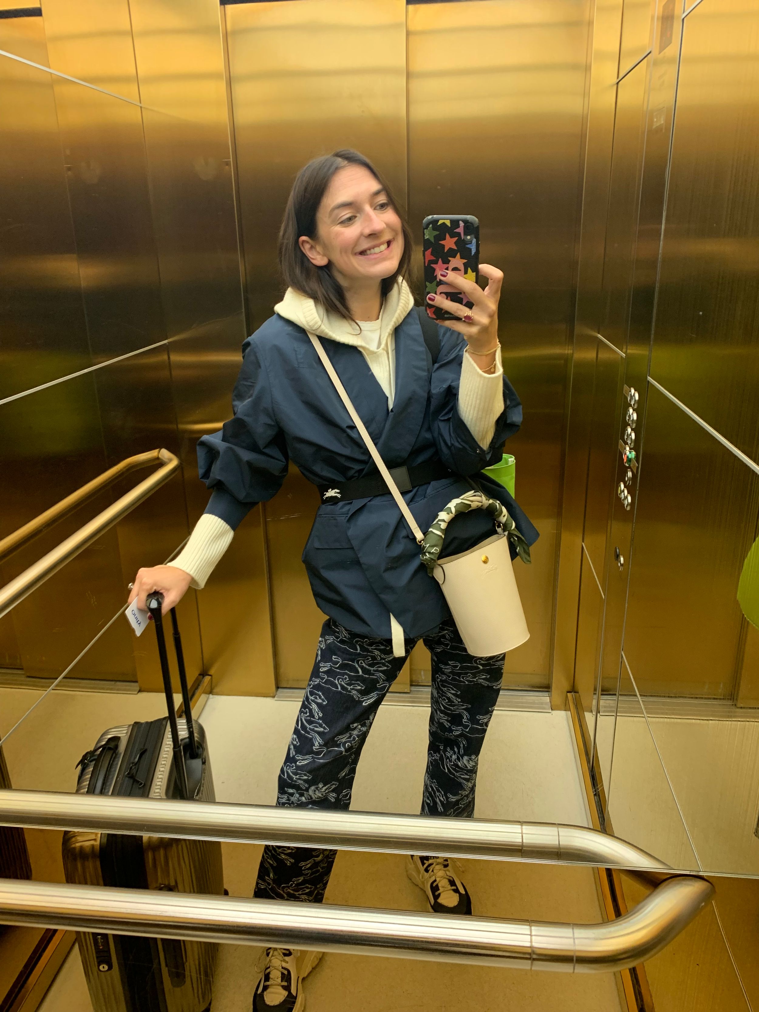 Juliette embraces travel chic in printed trousers, a belted navy kimono jacket over a cream hoodie, sporty sneakers, and a structured white bucket bag, ready for her next adventure.​