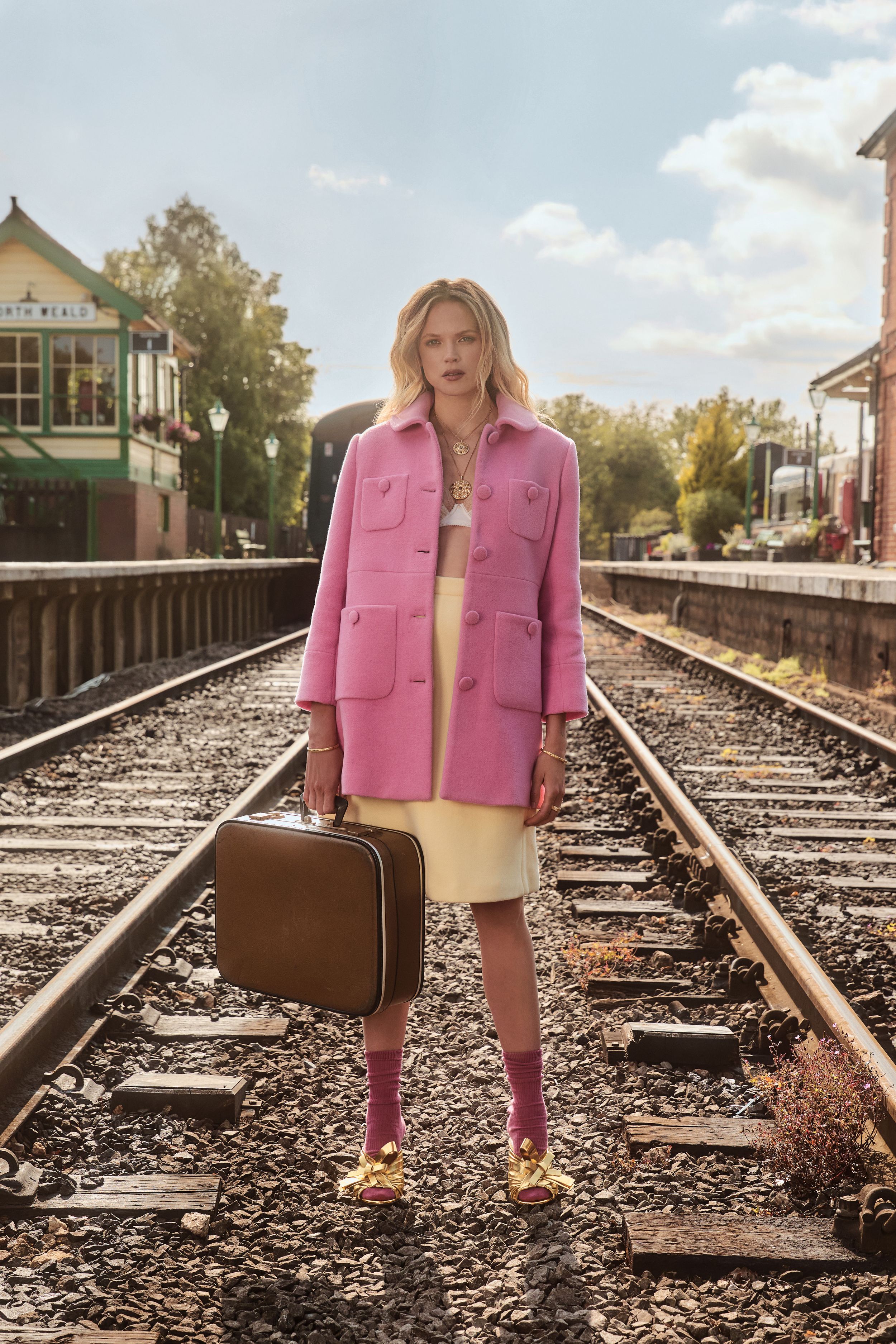 Dressed in a bubblegum pink coat and lemon-yellow skirt, Gabriella Wilde stands between railway tracks, clutching a vintage suitcase with gold bow heels and socks for a retro-modern statement look.