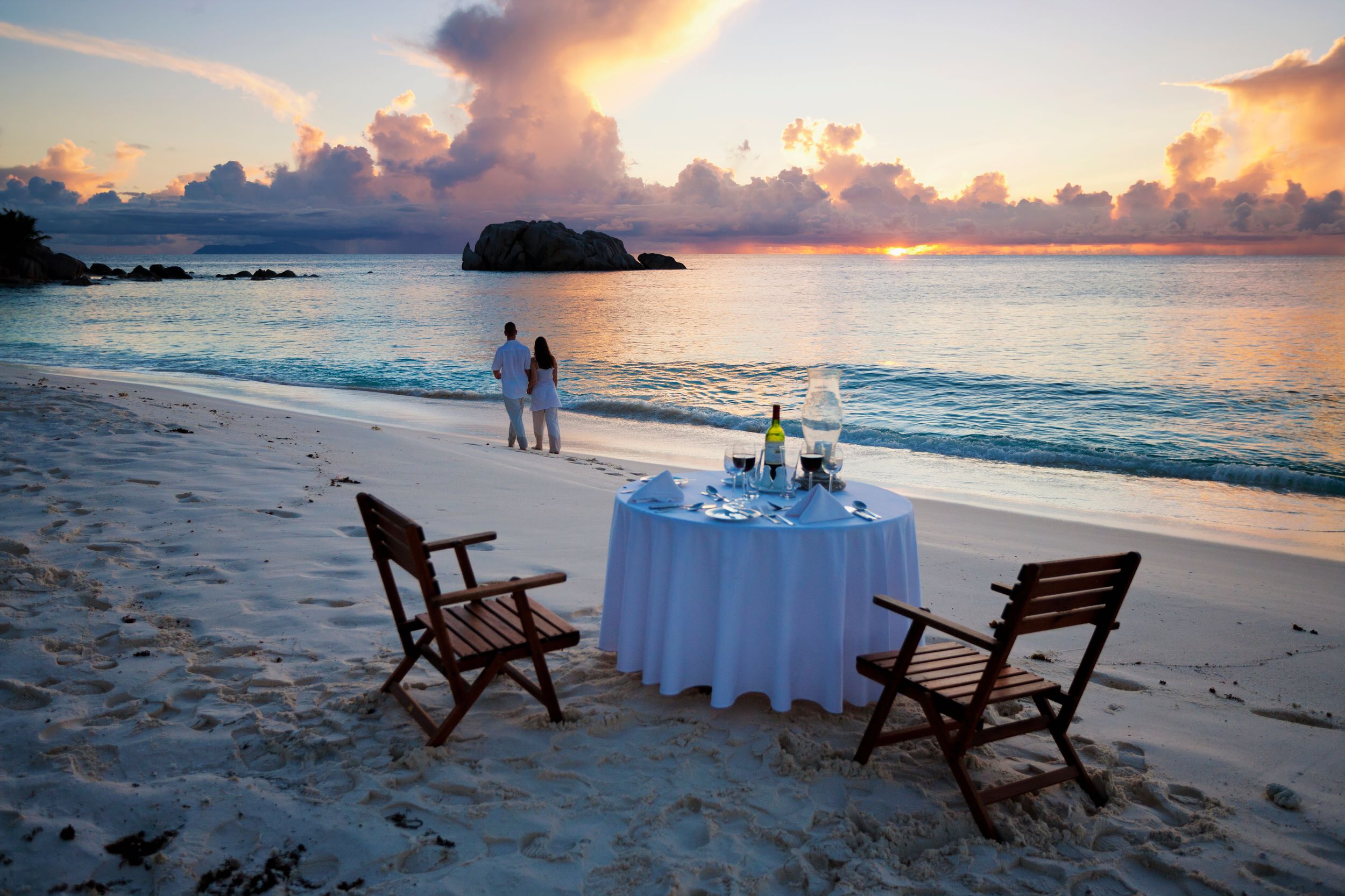 Romantic couple walking at sunset on beach with dinner table in foreground.Cousine Island.Seychelles