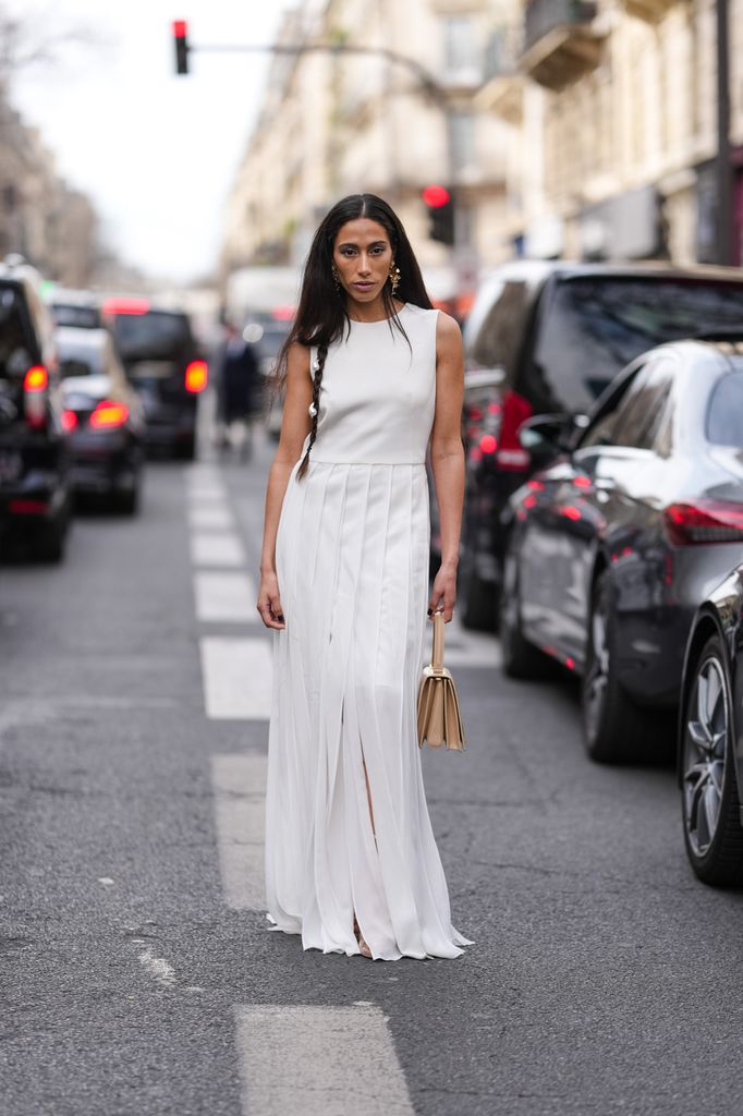 Ciinderella Balthazar wears braided hair with pearl detail, gold earrings, white sleeveless pleaded sheer mesh Akris maxi dress, light brown Akris leather bag, outside Akris, during the Paris Fashion week 