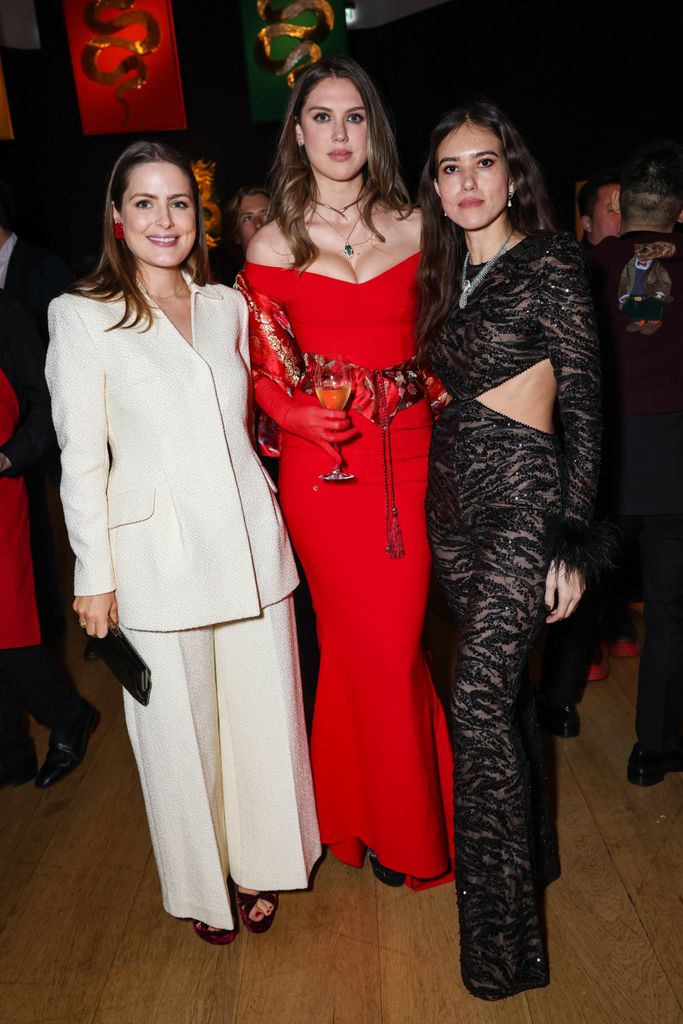 Philippa Cadogan, Lady Sabrina Percy and Natalie Salmon pose in front of hanging snake-themed banners. One wears a crisp white suit, another stuns in a bold red gown with a matching shawl, and the third dazzles in a black lace cutout dress with feather cuffs.