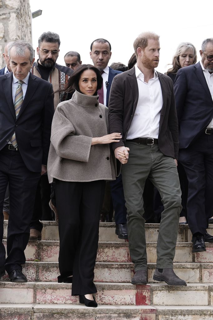Meghan en veste à col cheminée taupe descendant les marches avec le prince Harry avec des gens derrière eux