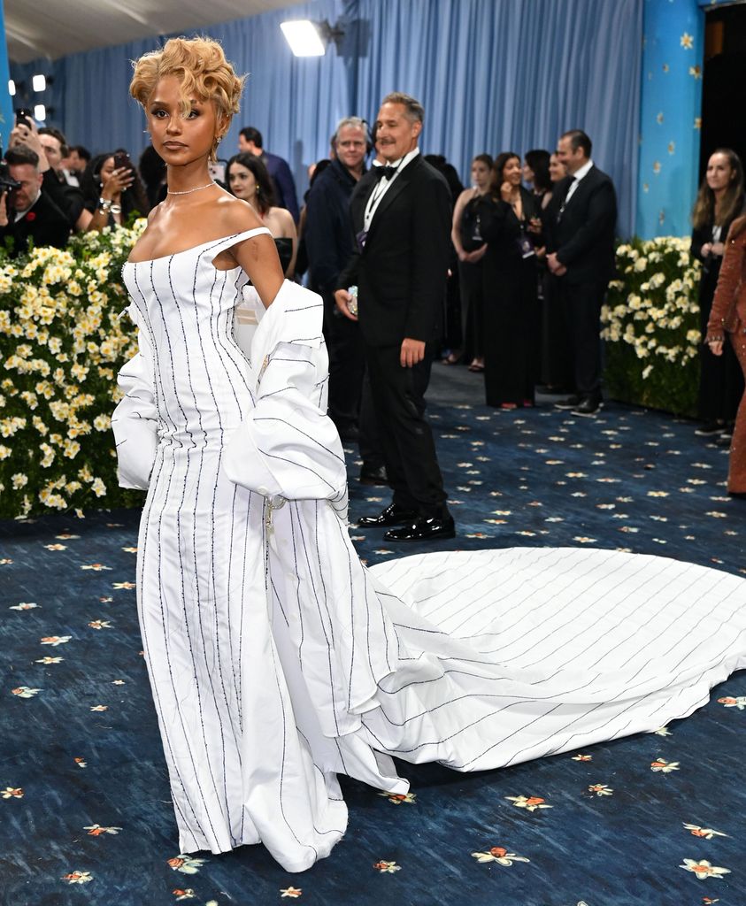 Tyla wears a white off the shoulder long dress with black stripe detail and a matching cape jacket. Her blonde hair is in a curly updo.