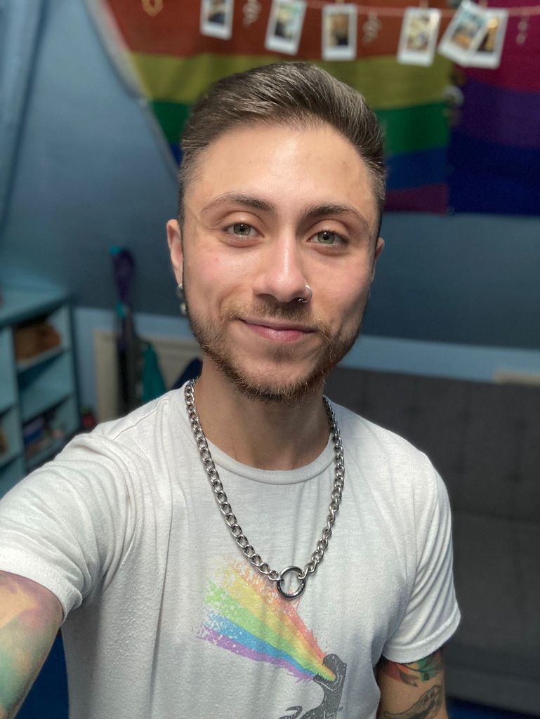 A man in a white t-shirt that features a T-rex spitting out rainbow. He also wears a chain necklace. Behind him are a pride flag and bisexual pride flag on the wall