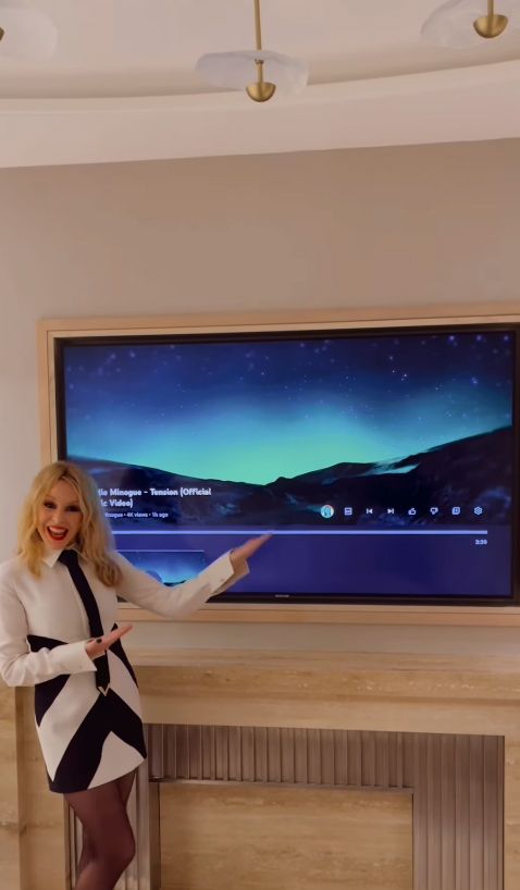 Kylie Minogue in a mini dress gesturing at a flat screen TV