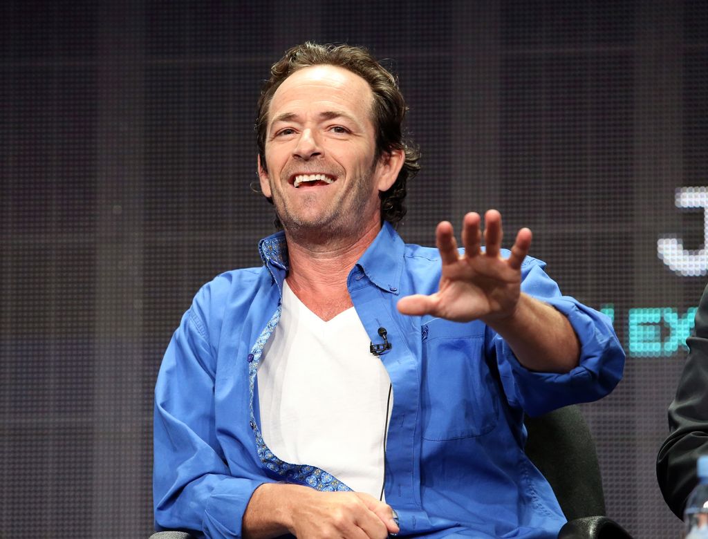 Luke Perry tend la main en parlant sur scène.
