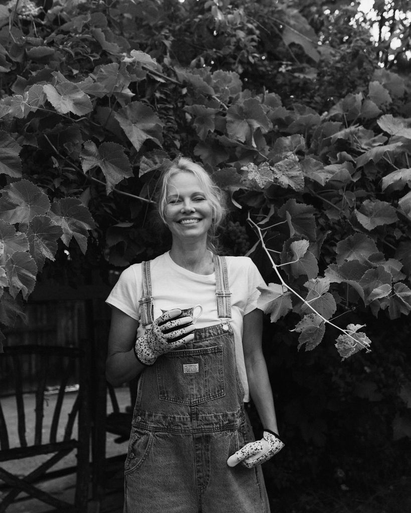 Pamela Anderson in her denim dungarees and white t-shirt stands in her garden holding a mug. She is also wearing gardening gloves. 