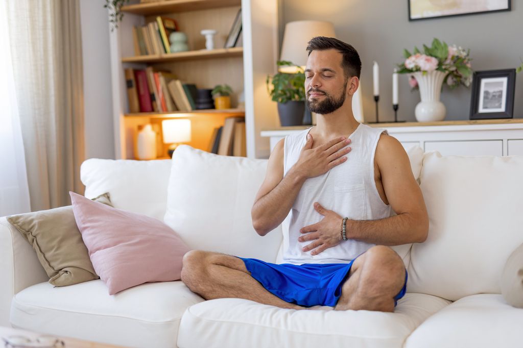 A man in white tank top and blue shorts sits on a white couch in his living room with hands on his chest, reflecting peacefully in an environment filled with calm and warmth