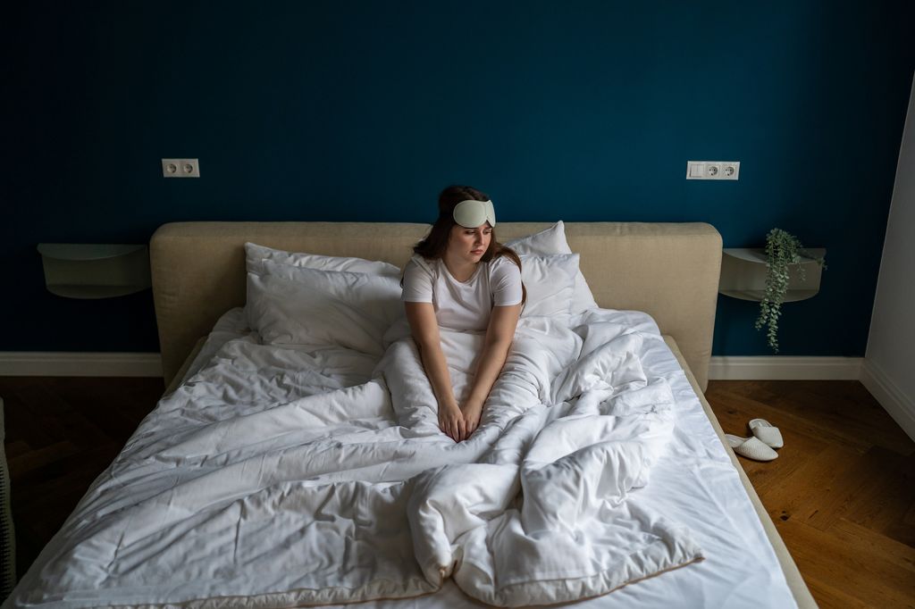 Exhausted unhappy young woman wearing sleep eye mask sitting in bed feeling tired and depressed after wake up in morning. Depressed female suffering of insomnia. Sleep deprivation and depression