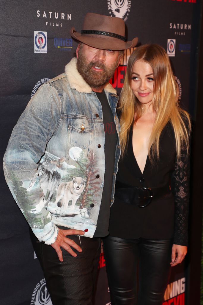 Nicolas Cage in denim jacket with wolves on it and a cowboy hat standing with Erika Koike