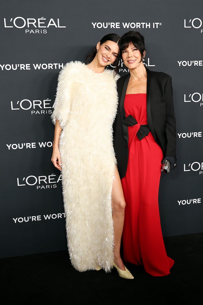  Kendall Jenner and Kris Jenner stand on the red carpet embracing. Kendall wears a textured cream dress and Kris a red dress with black blazer
