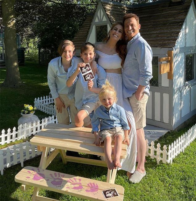 a family of three boys and a father and pregnant mother smile at the camera as they pose for a family photo in front of a wendy house on a sunny day holding a sonogram