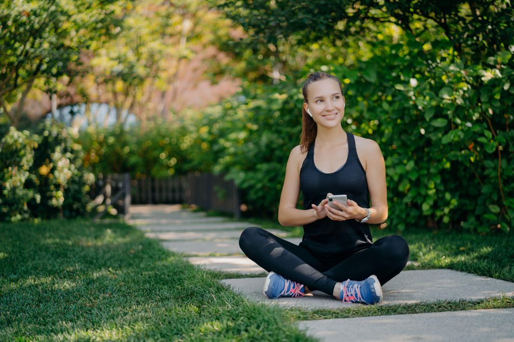 Sportswoman sits crossed legs outdoors checks newsfeed via smartphone
