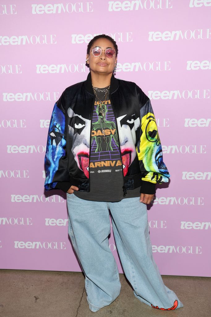 Raven Symone with short black hair, wearing a colourful jacket, jeans and pink tinted sunglasses