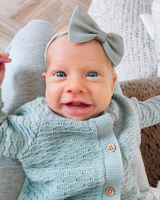 a very young baby wearing a blue cardigan and matching blue bow headband looks at the camera and smiles with her gums showing and big blue eyes open wide