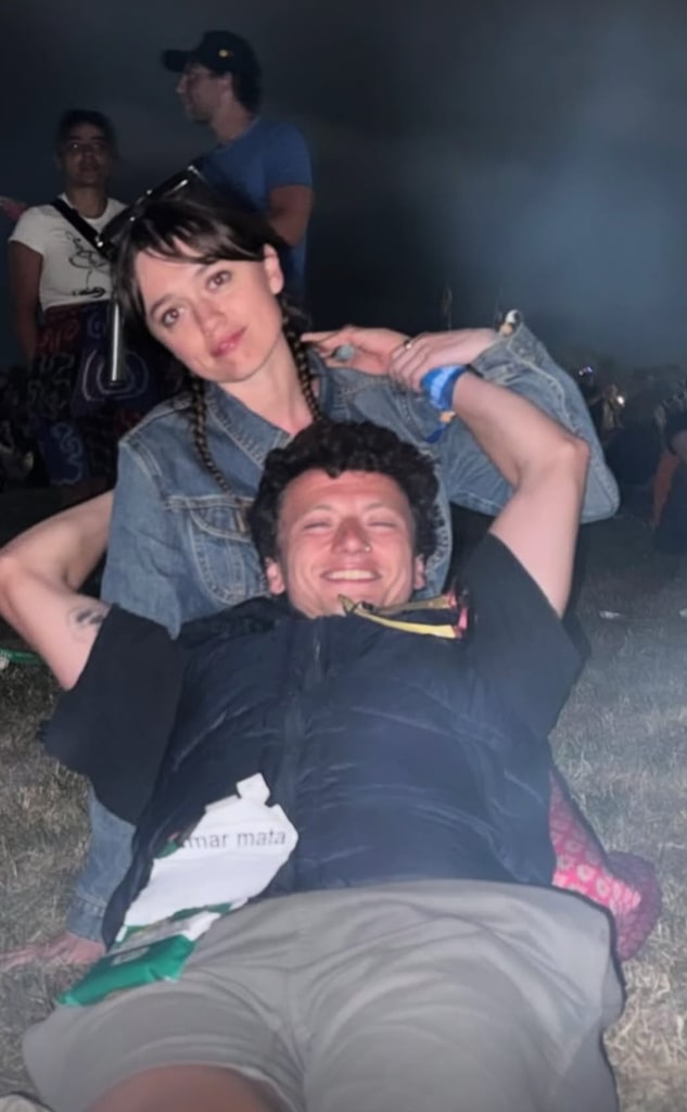 In this candid, warmly lit photograph taken at an outdoor evening music festival, actress Aimee Lou Wood is sitting on the grass with actor Adam Long resting his head playfully on her lap. Aimee exudes casual yet effortless style in a light denim jacket with her hair parted down the center and braided in pigtails, giving her look a mix of youthful charm and bohemian polish. A pair of dark sunglasses rests on her head like a crown. Adam, clearly relaxed and beaming with joy, is wearing a black vest and light-colored pants, his body stretched across the grass in front of her. They are surrounded by other festival-goers, some standing, some seated, but the focus is on the couple’s easy intimacy and carefree energy. The setting is dusky, likely late evening, with the faint haze of stage lights and smoke lingering in the background.