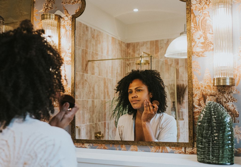 A beautiful young black woman applies foundation / concealer with a make-up brush in a bathroom mirror.