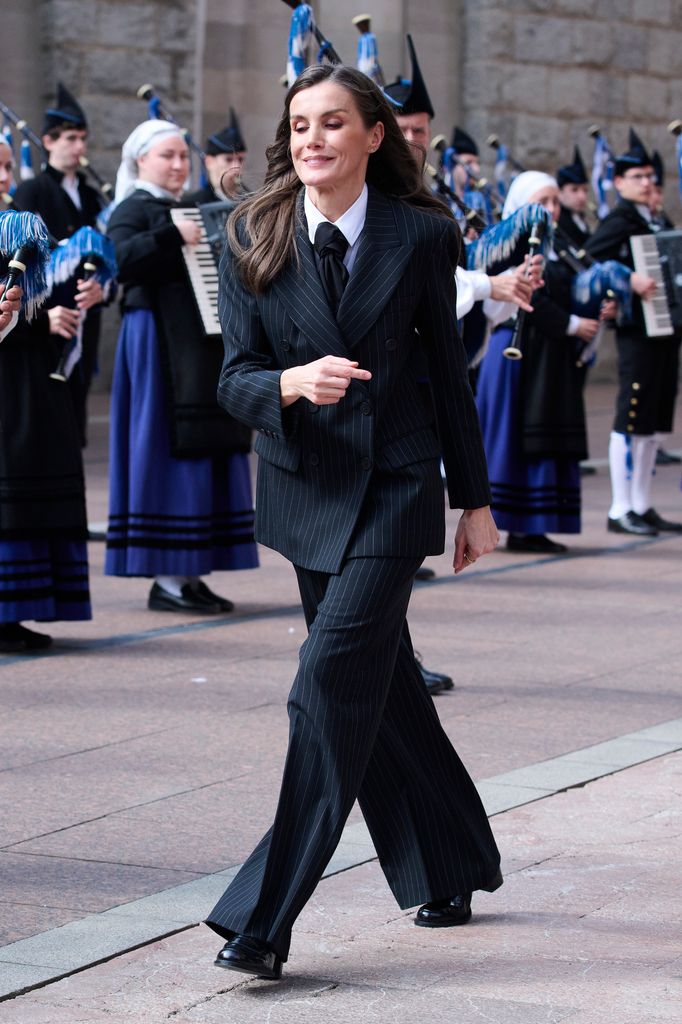 Queen Letizia of Spain walking out wearing a black pinstripe suit, loafers and a black tie