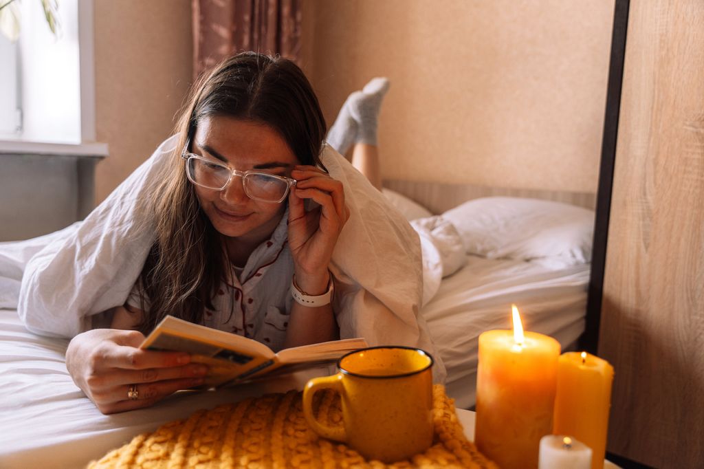 Young woman in glasses for poor eyesight reading book with mug of hot drink and next to candles under blanket, relaxing cozycore.