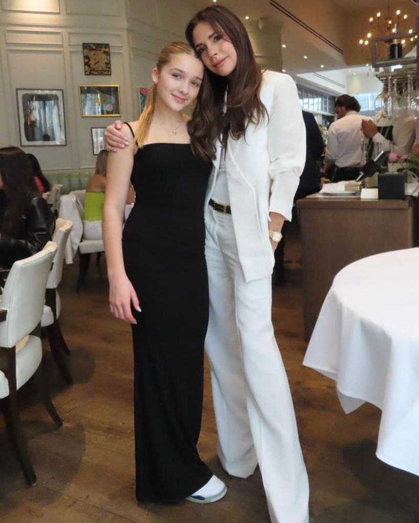 Harper Beckham in black dress posing with mother Victoria Beckham in white suit