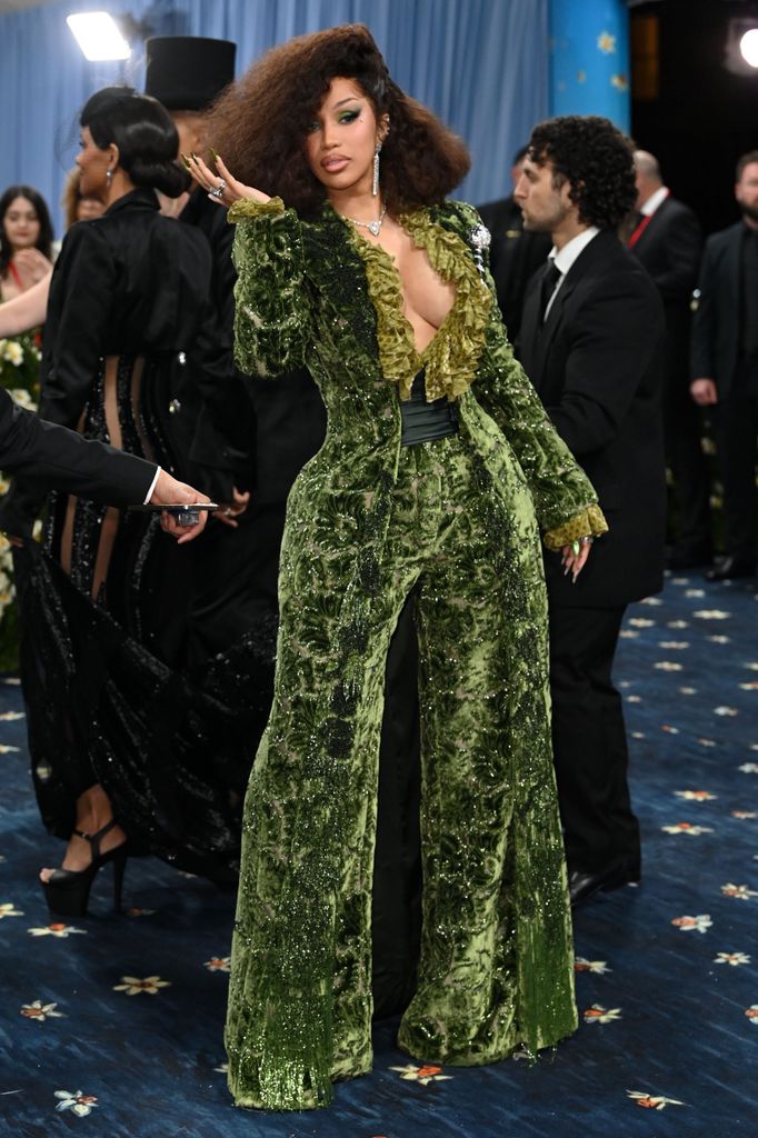 Cardi B wears a green velvet jacket and matching wide leg trousers. Underneath her jacket is a frilly green shirt.