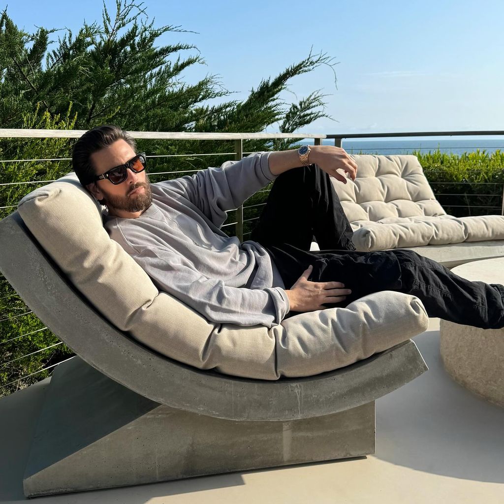 Scott Disick lying on an outdoor seat in the sun wearing a grey top