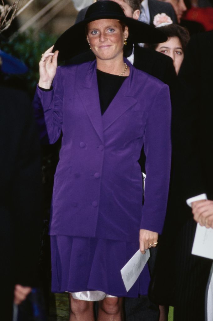Sarah, Duchess of York wearing a baby bump-skimming purple outfit and holding her hat at the wedding of the Marquis of Marlborough and Rebecca Few Brown