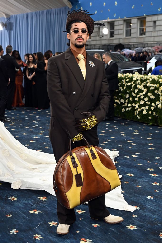 Bad Bunny wears a black suit with brown shirt andy ellow tie. He wears patterned gloves, an intricate hat and carries a large brown and yellow bag.