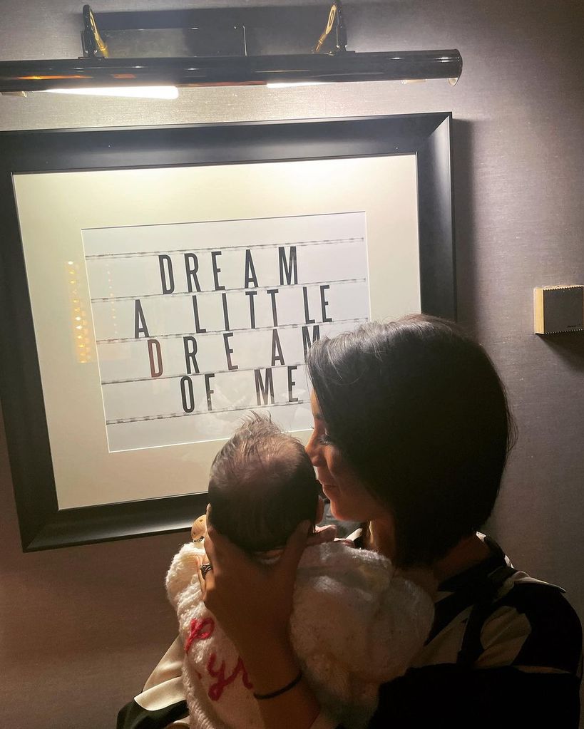Janette Manrara and her baby Lyra in front of a wall hanging 