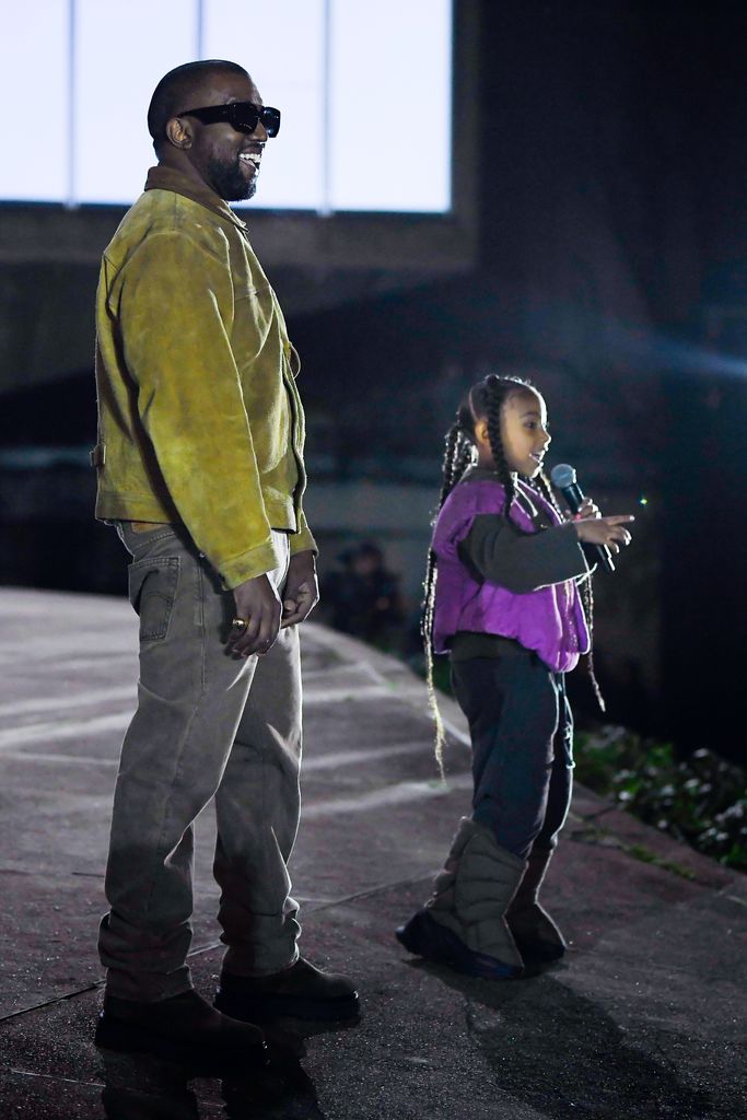 North on stage holding a mic and gesturing to the crowd as she raps, her father Kanye is stood behind her