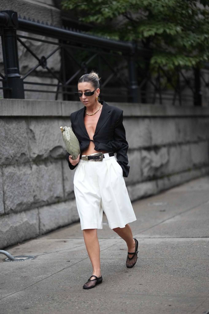 A guest wears sunglasses, a black oversized blazer jacket, a belt, white shorts, mesh shoes, a green bag, outside Khaite , during New York Fashion Week, 