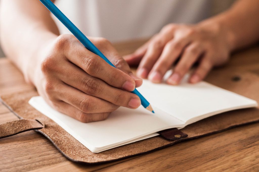 Close-up view of a man writing on a leather-bound journal with blue pencil