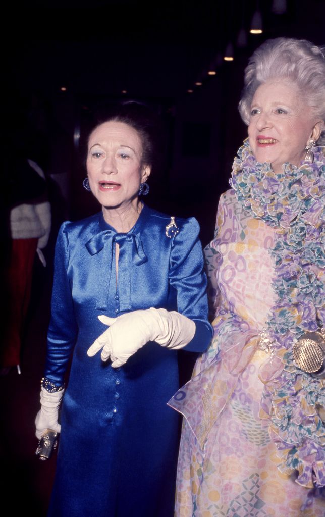 Wallis Simpson wears a royal blue satin gown with white opera gloves and statement jewelry, standing beside Mildred Hilson in a pastel ruffled dress at the 1974 Met Gala.