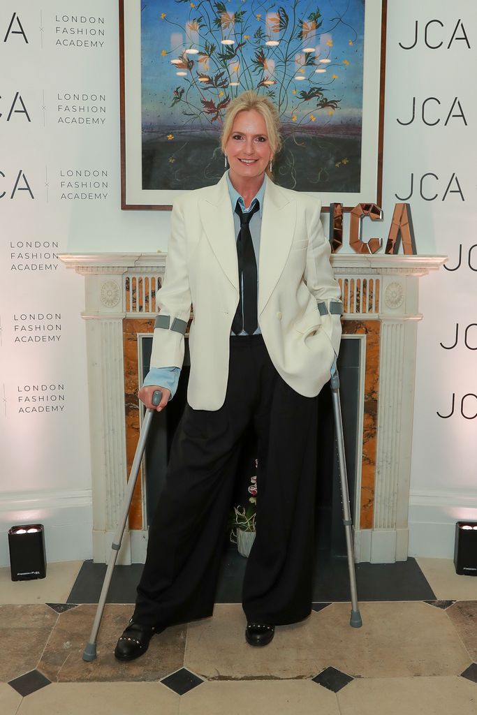 Penny Lancaster at event on crutches in black and white suit with tie