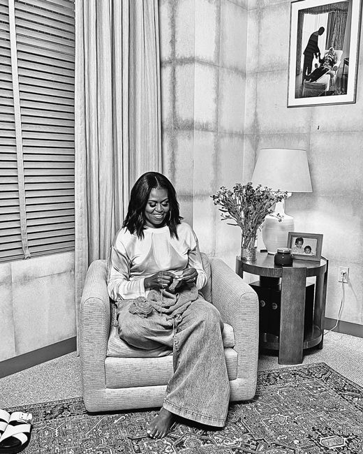 a black and white image of michelle curled up on an armchair in a high ceilinged llight coloured room with a large window as she knits beside a table featuring a family photo