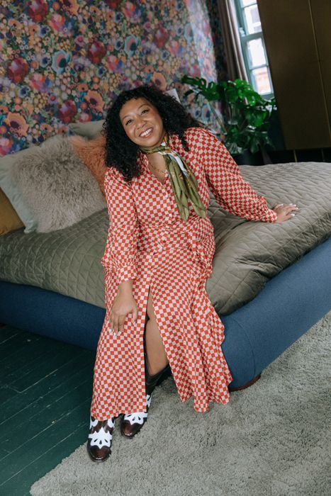 Nicole Ocran wearing a check print long sleeved dress, smiling sitting on a bed