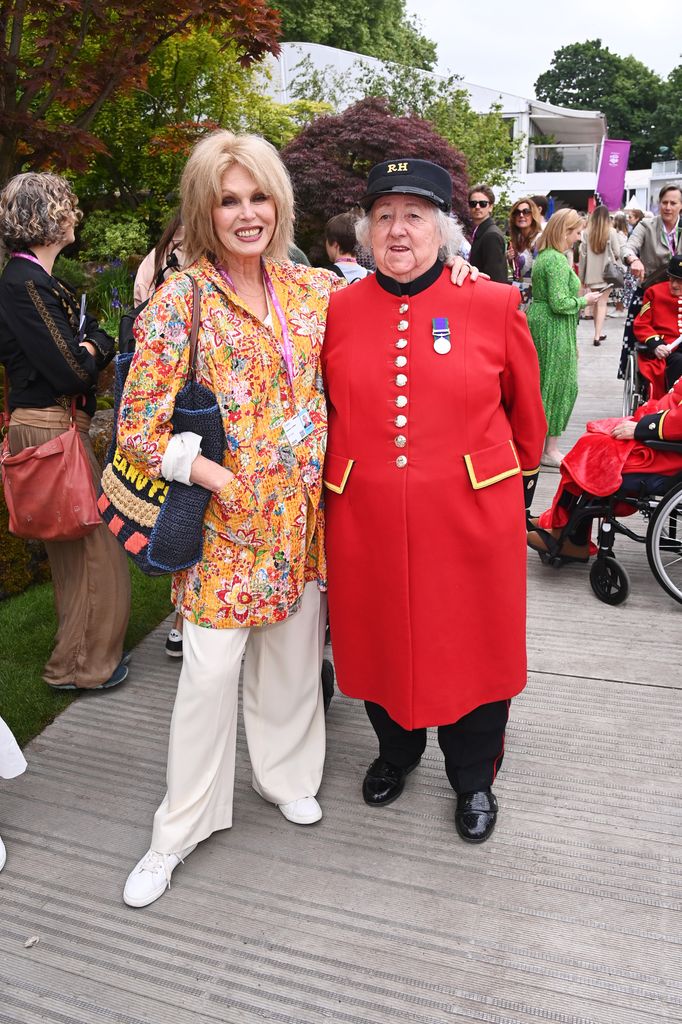 Joanna Lumley wears a vibrant orange floral jacket and cream trousers, standing next to a Chelsea Pensioner in a red uniform.