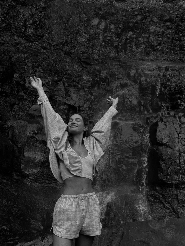 Lily James poses in front of a waterfall in a black and white snap, her hands in the air and she smiles with her eyes closed. She wears a checked cropped shirt and matching boxer shorts. 