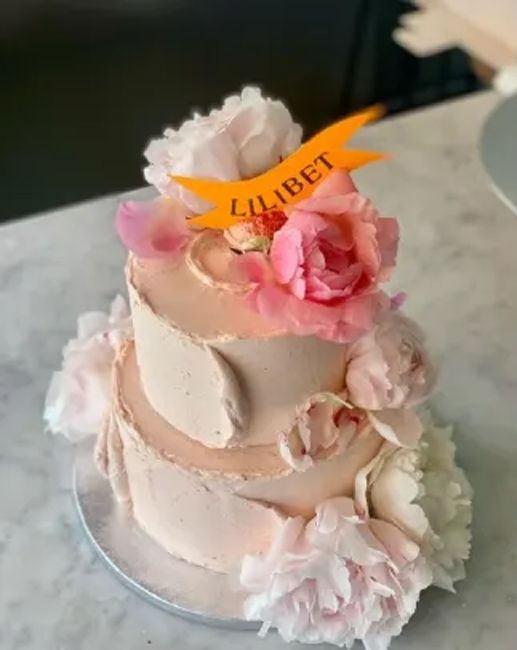 a delicate looking two tiered cake stands with light pink buttercream roughly smoothed all over it with blush hued peony flowers stuffed around the edges and an orange banner at the top says lilibet