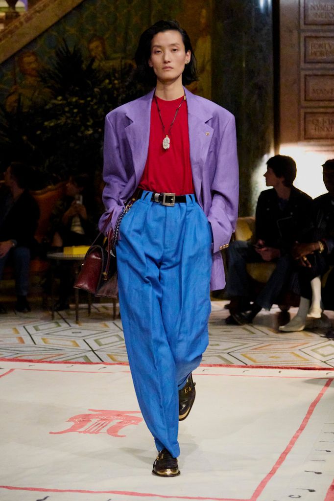 A model wears bright blue high waisted trousers, a lilac blazer with slouching shoulders and a red top