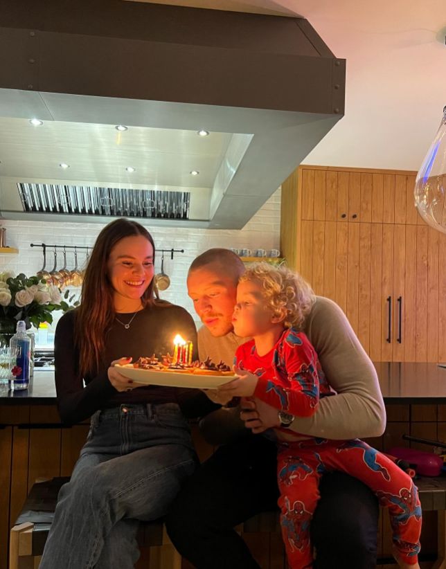 Holly Ramsay holding a birthday cake while Adam Peaty and his son blow out the candles