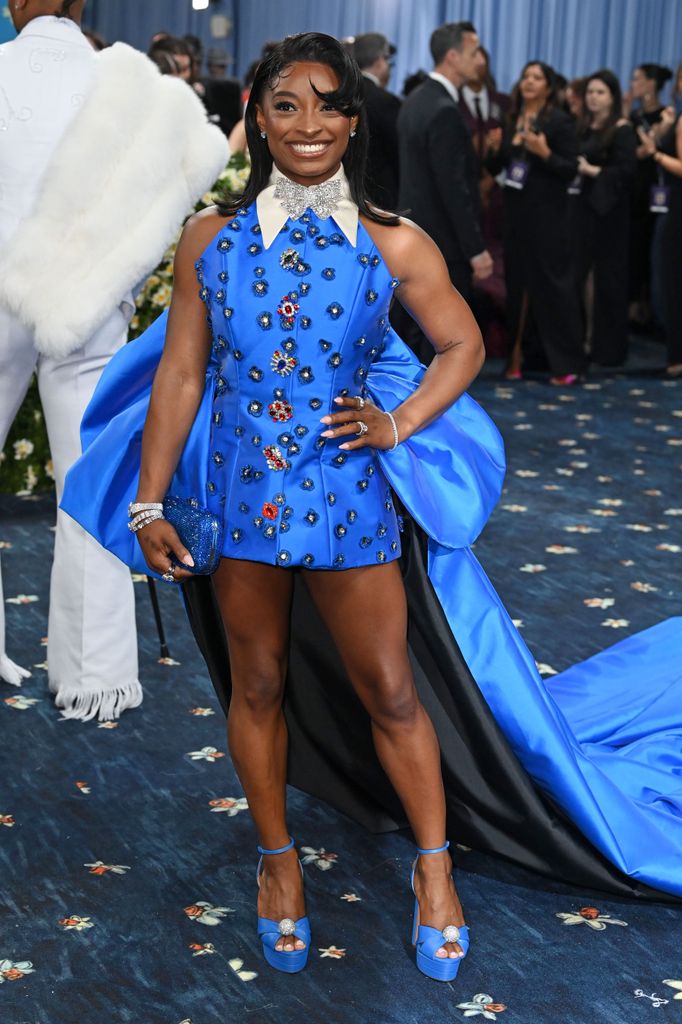 Simone Biles wears a bright blue mini derss with blue platform heels and a big bow train at the back