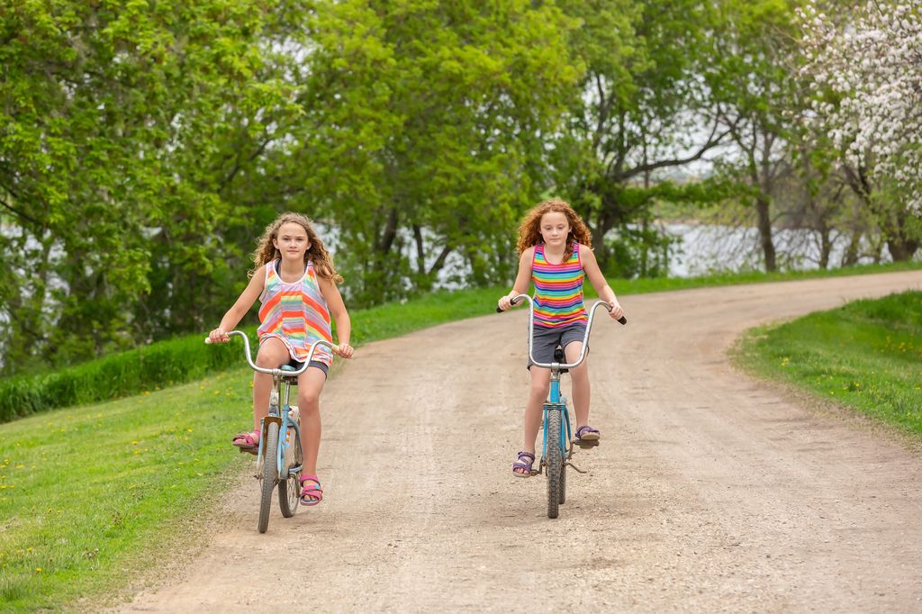 Two young girls (sisters) riding old, rusty, vintage bikes down a rural gravel driveway.