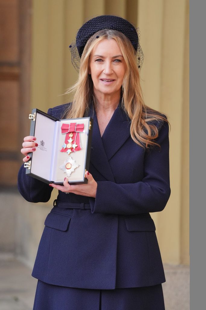 Anya Hindmarch, in a navy coat and hat, smiles while holding an OBE medal inside an open black case.