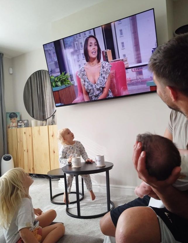 Aljaz Skorjanec holding a baby as they and two other children watch Janette Manrara on TV