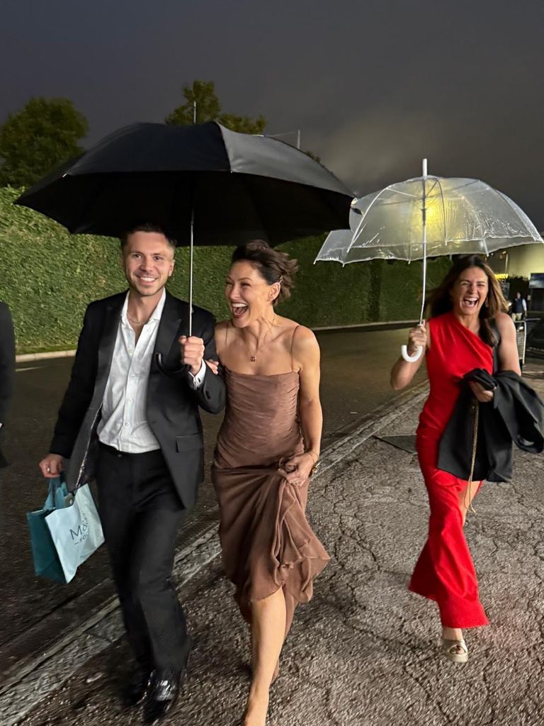 Emma Willis wearing a brown dress and walking under a black umbrella