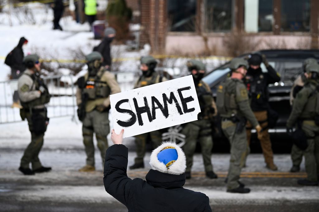 An onlooker holds a sign that reads "Shame" as members of law enforcement work the scene following a suspected shooting by an ICE agent 