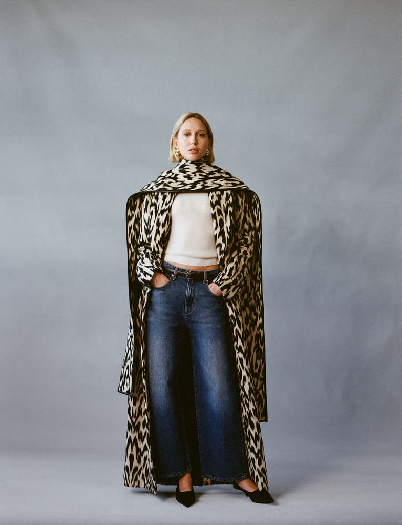 Princess Olympia of Greece stands against a grey backdrop wearing a striking patterned coat with a matching scarf draped over her shoulders. She pairs the ensemble with a white crop top, high-waisted wide-leg blue jeans, and black pointed-toe heels, completing the look with gold earrings and a sleek bob.