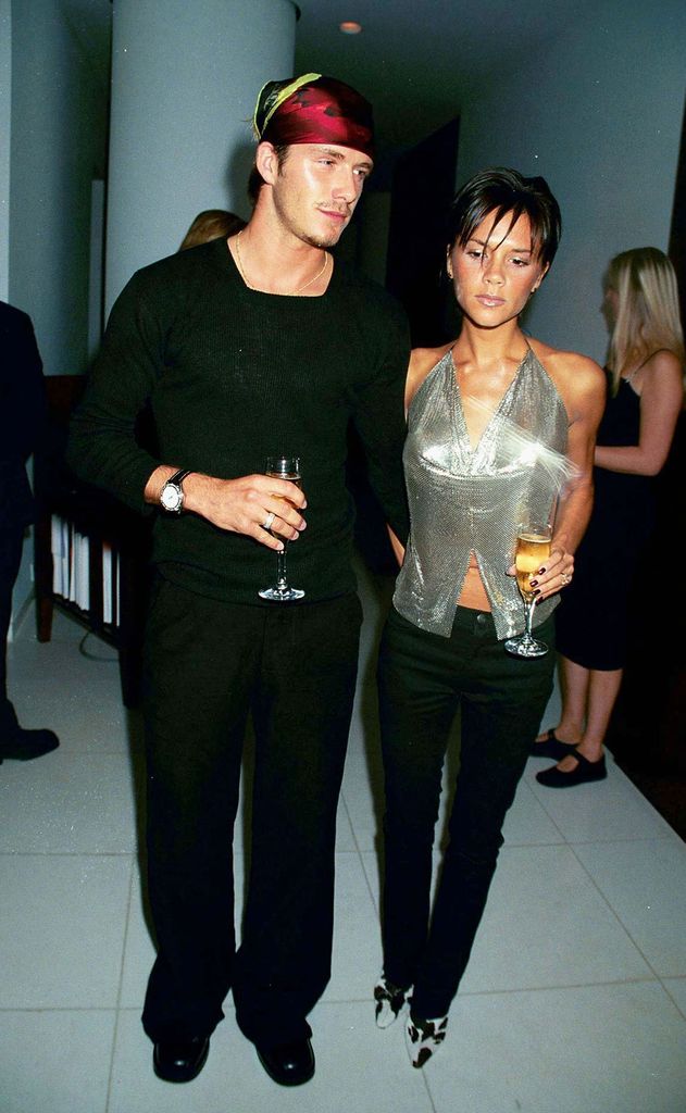 Victoria Beckham wears a shimmering silver chainmail halter top with black trousers and cow-print heels, standing next to David Beckham in all black at a 1999 fashion party.