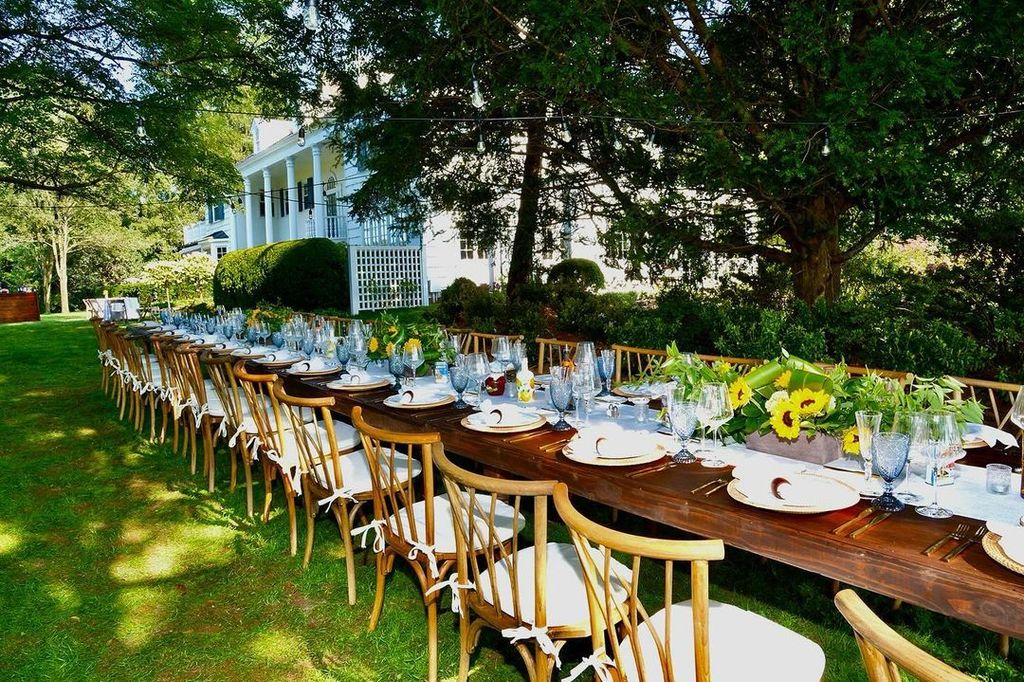 A long oak table covered in white plates and sunflowers sits in a garden